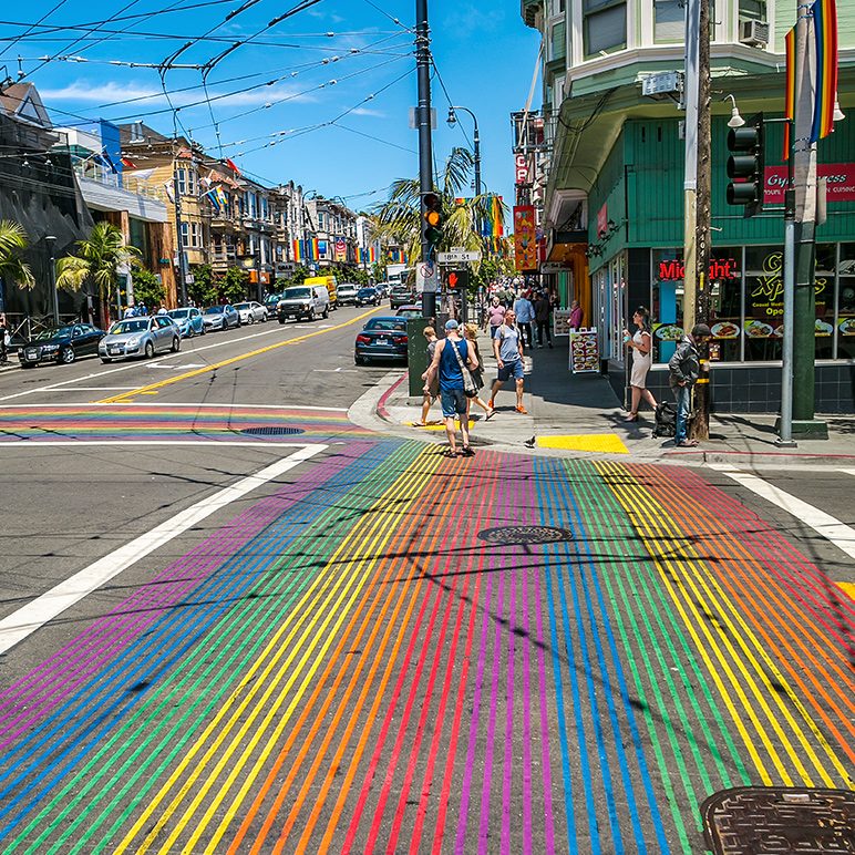 Rainbow-striped crosswalk on a busy city street with pedestrians and colorful buildings on a sunny day.