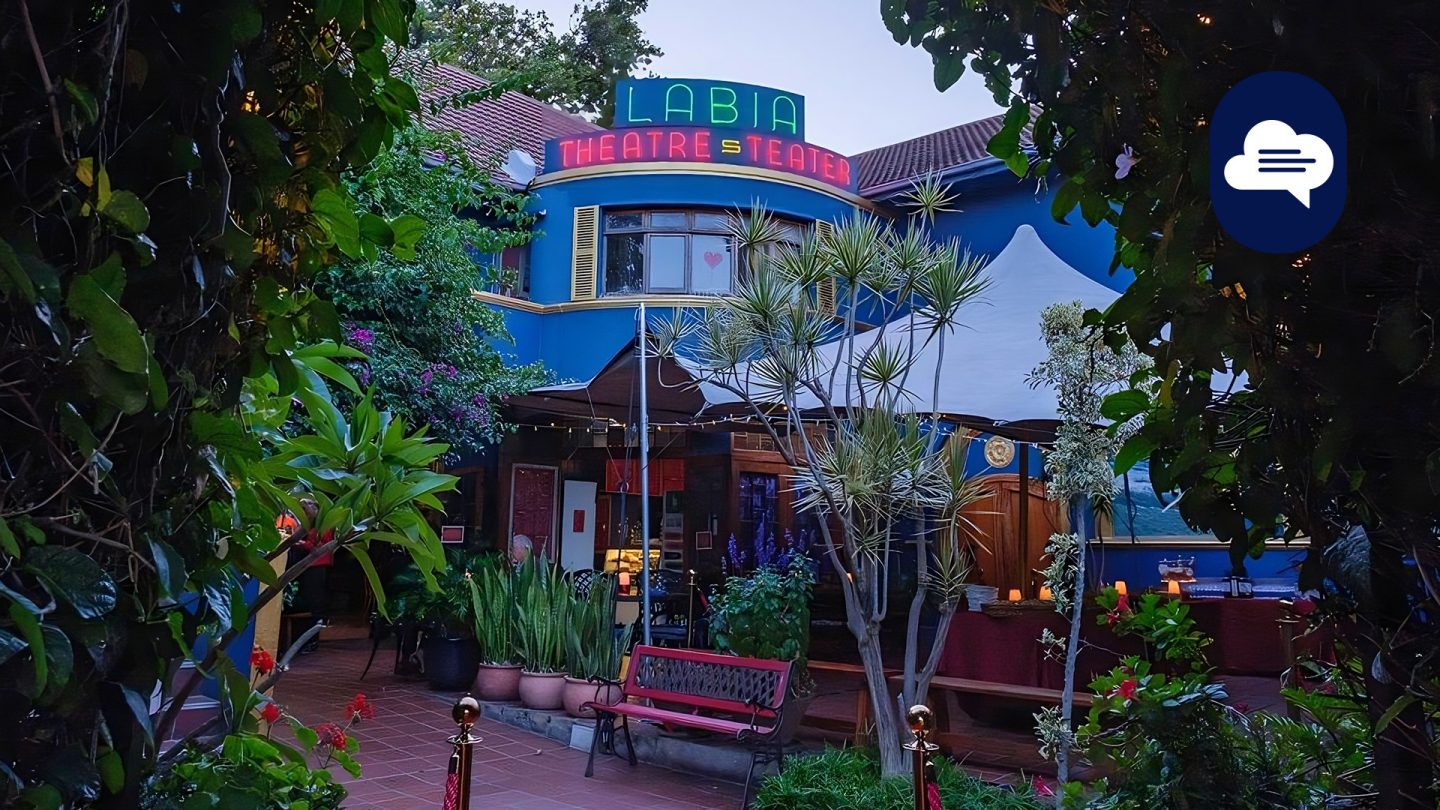 Exterior of the Labia Theatre with blue walls, neon sign, and surrounded by green plants