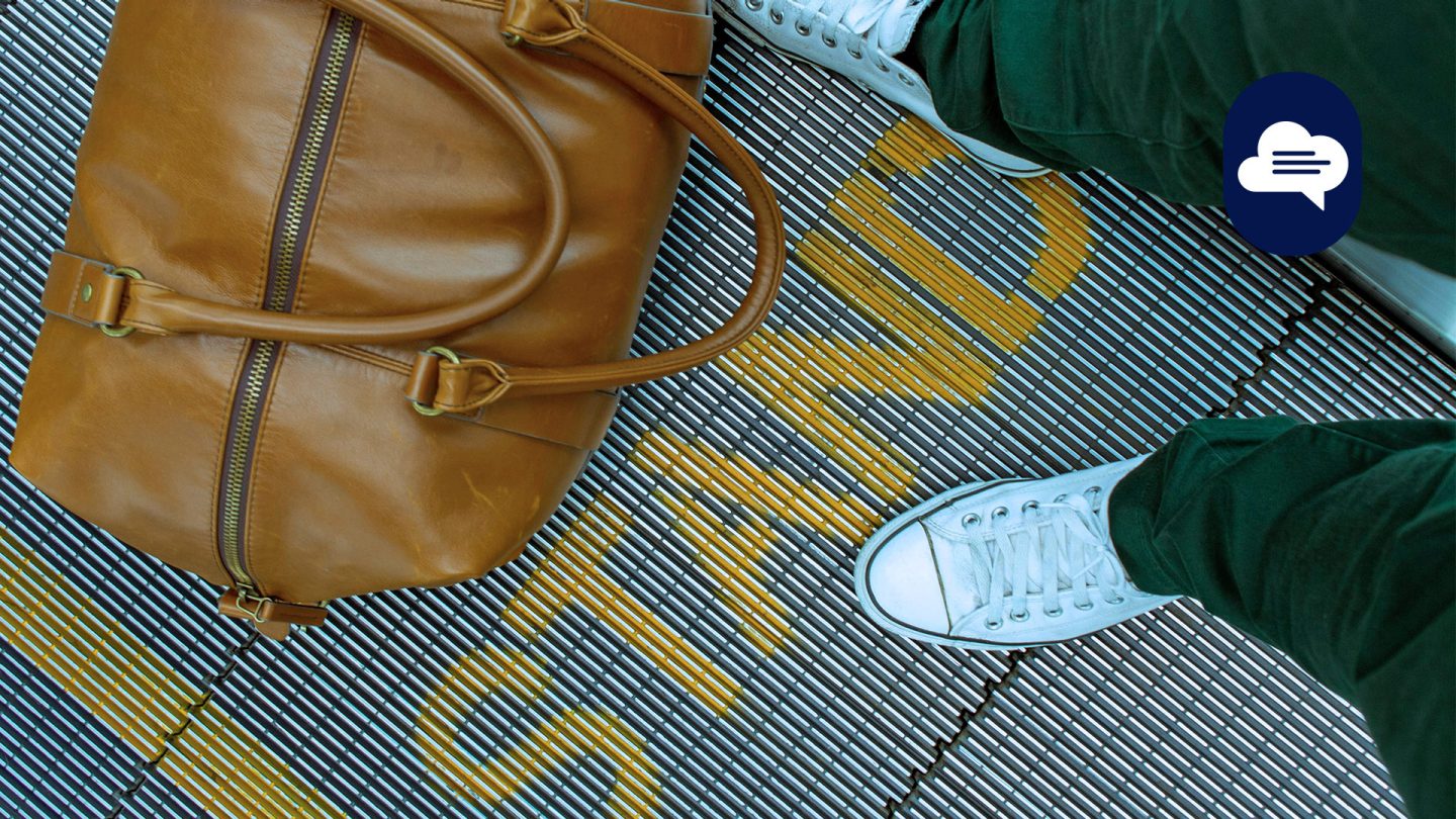 Team Stories: Blick von oben auf die weißen Turnschuhe einer Person, die auf einem Fahrsteig neben einer Reisetasche aus braunem Leder steht.