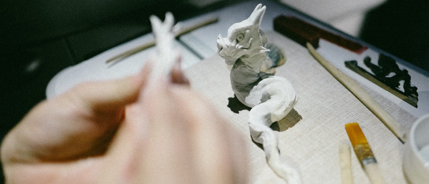 Close-up of hands sculpting a small dragon figure from white modeling clay