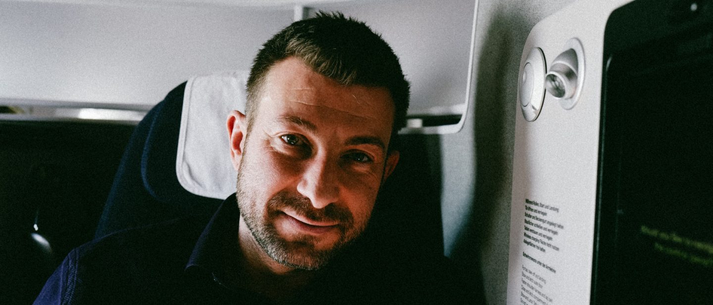 Man with short hair and beard smiling while sitting in an airplane seat next to a window and reading light.