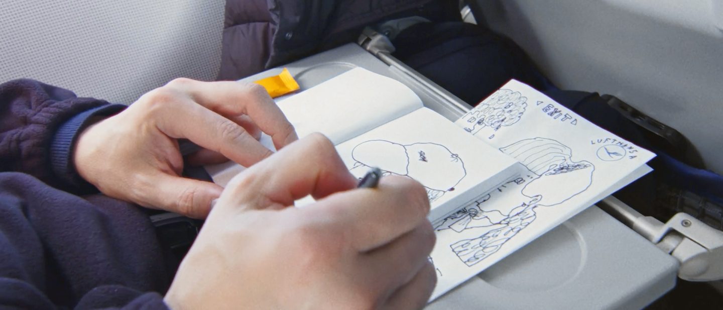 Hands sketching drawings in a notebook with Lufthansa logo while on an airplane