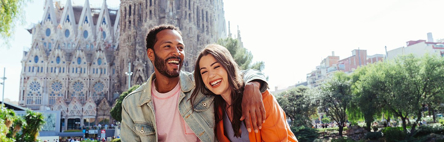 A young couple in Barcelona stands smiling in front of the Sagrada Familia.