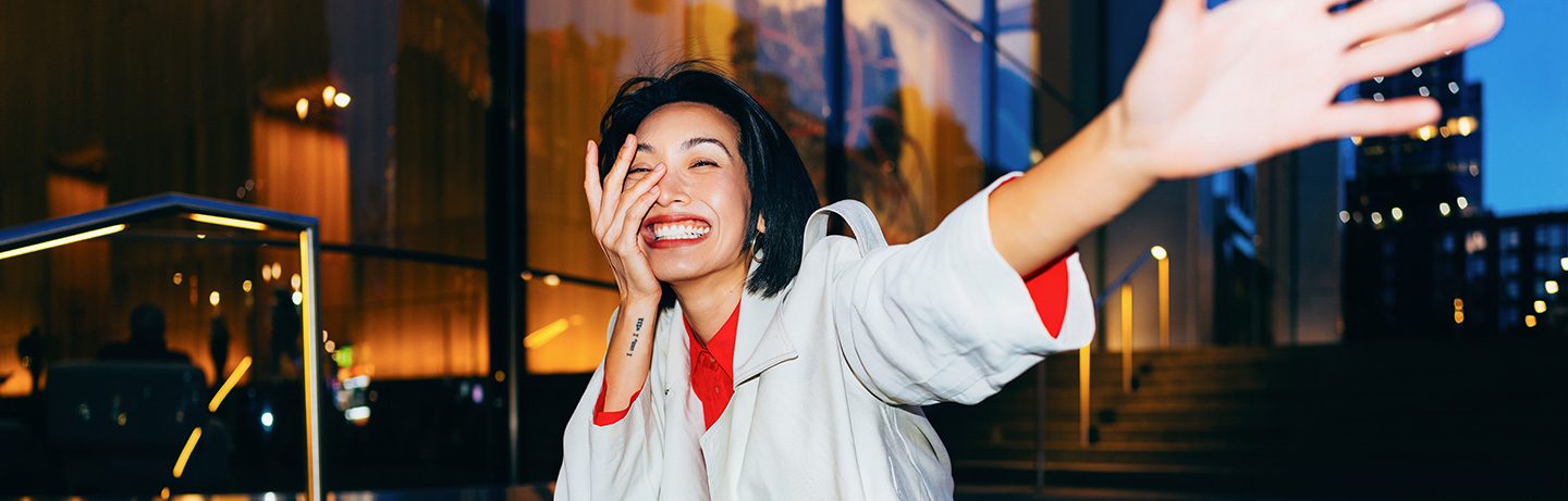 Laughing woman in front of a lit building raises one arm in the air.