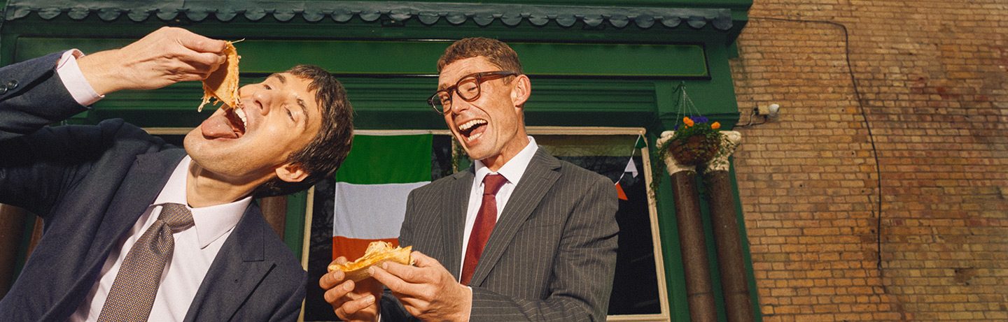 Two young men enthusiastically eat a pizza outside an Irish pub.