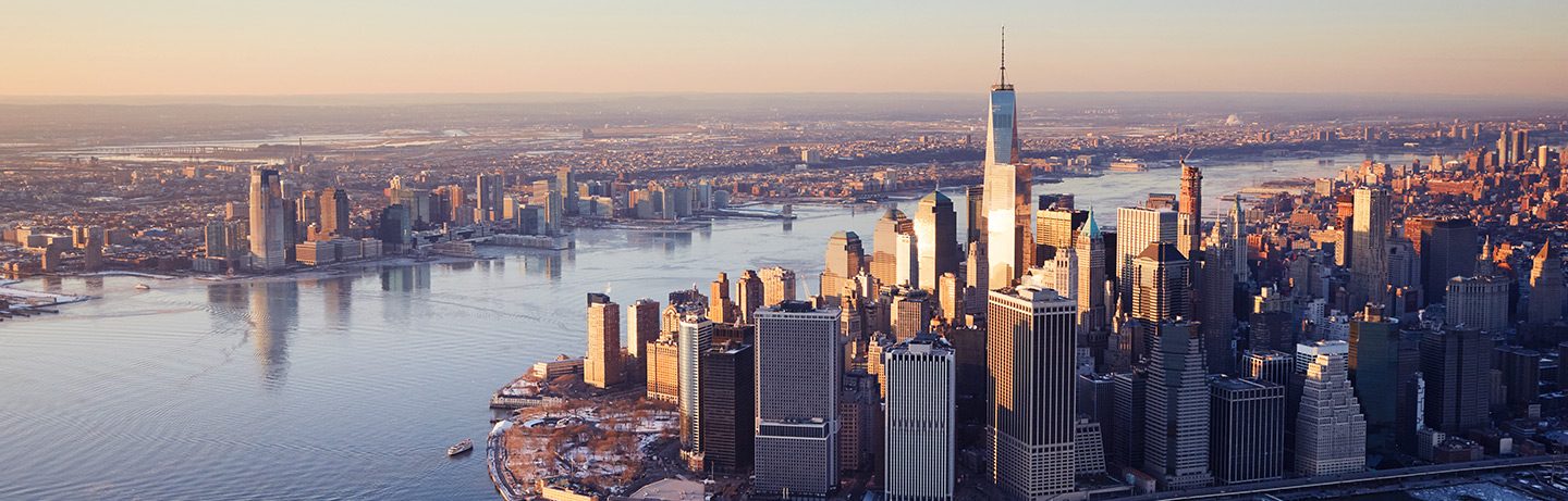 Skyline of skyscrapers along a waterfront, featuring a prominent glass tower in warm evening light.