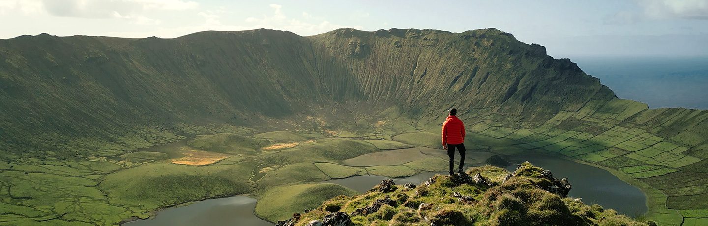 A person in a red jacket overlooks the crater lake in a wide green landscape