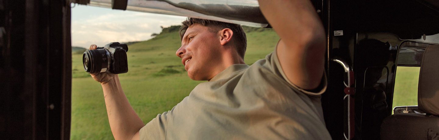 Man on safari taking photos of a grassy savanna through the open roof of his game drive vehicle.
