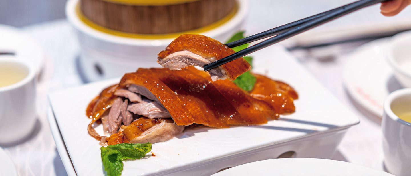 Mit Stäbchen wird ein Stück knusprige Ente von einer Servierplatte gehoben.