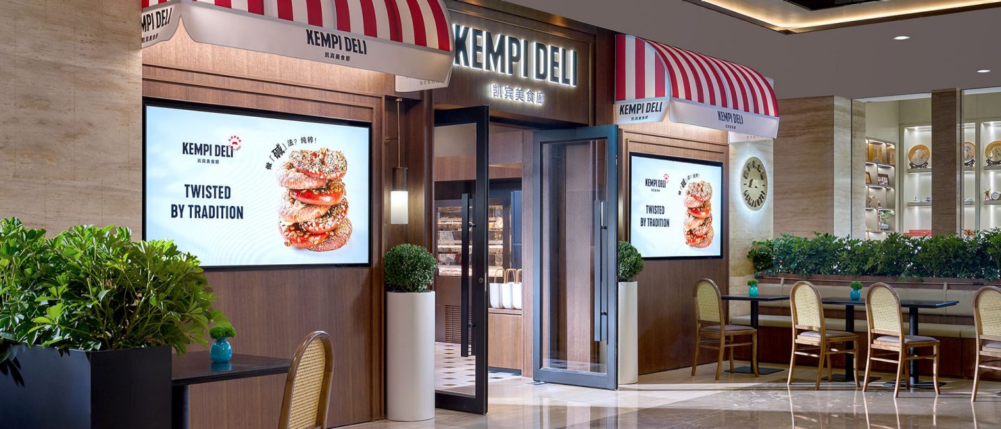 Eingang des Restaurants Kempi Deli mit rot-weiß gestreiften Markisen, beleuchteten Werbedisplays und Sitzplätzen im Innenbereich.