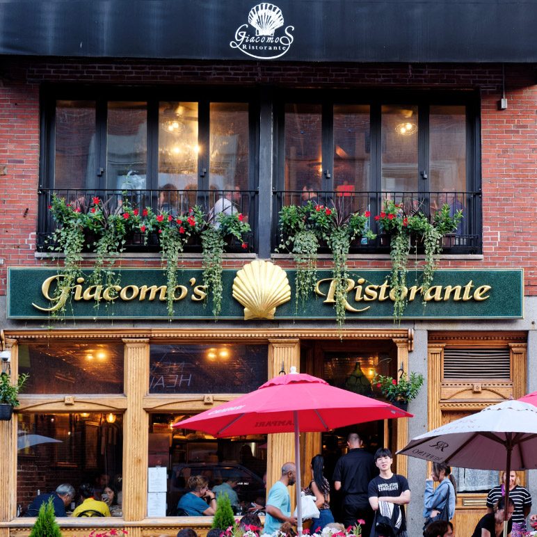 Die Fassade von Giacomo’s Ristorante in Boston mit Gästen und roten Sonnenschirmen