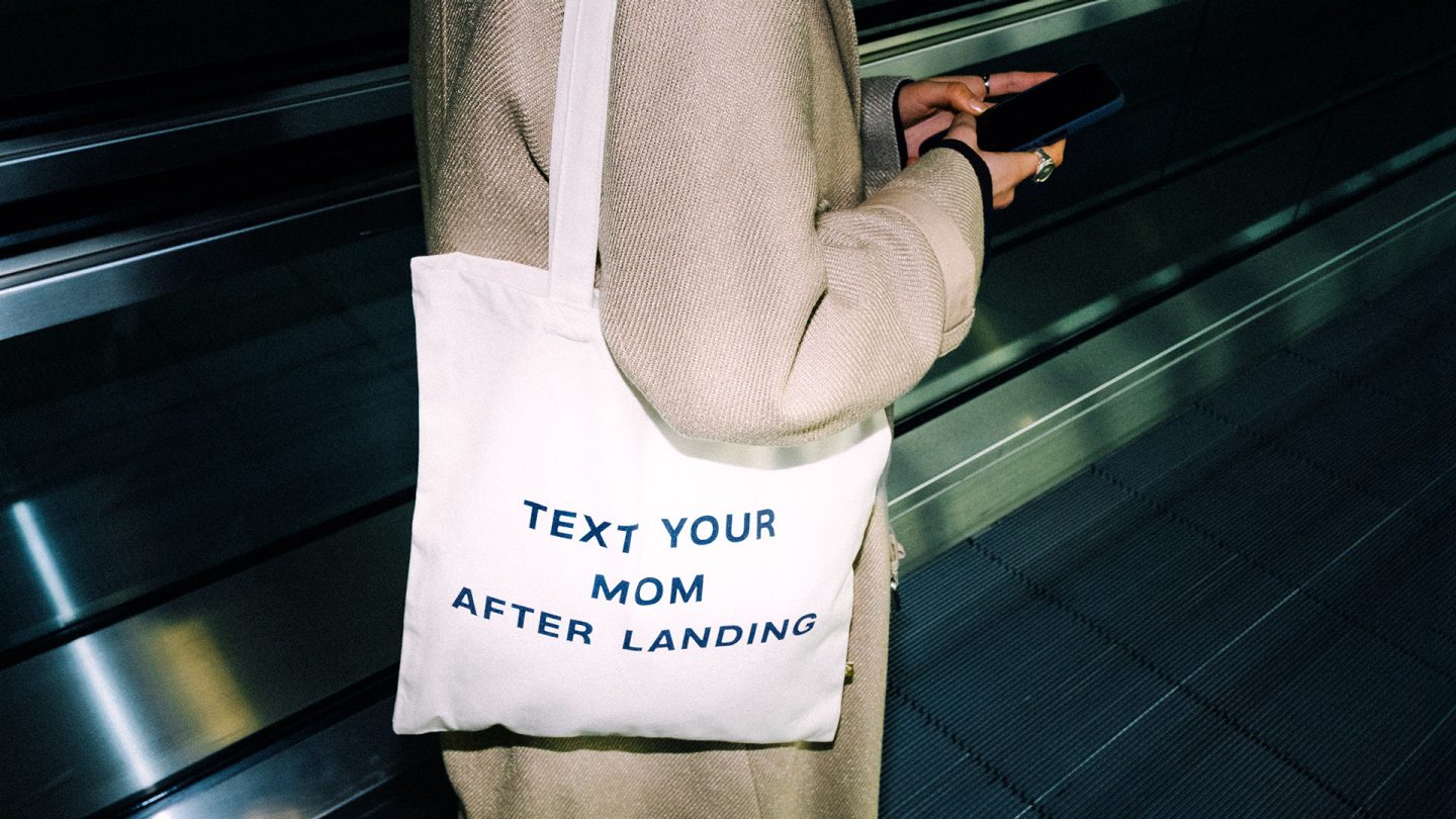 Weiße Stofftasche mit der Aufschrift „Text your mom after landing“ an einer Person auf einem Rollband im Flughafen