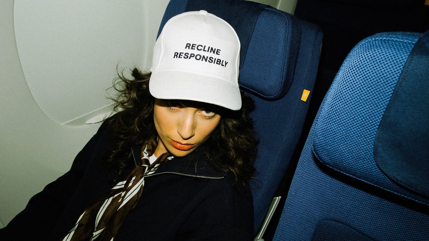 Person sitzt in einem Flugzeugsitz und trägt eine weiße Kappe mit der Aufschrift „Recline responsibly“