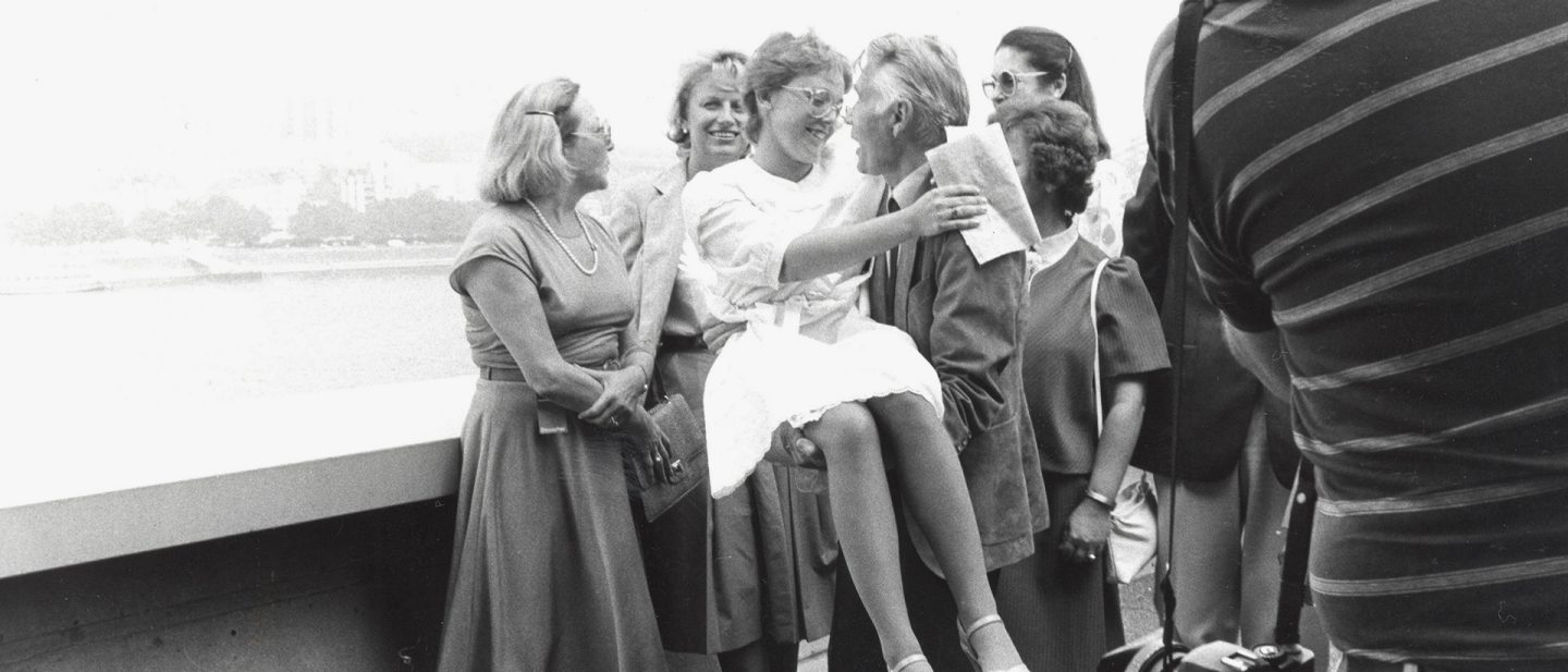 Historisches Schwarz-Weiß-Foto einer Gruppe von Menschen im Freien; ein Mann hebt ein junges Mädchen freudig hoch