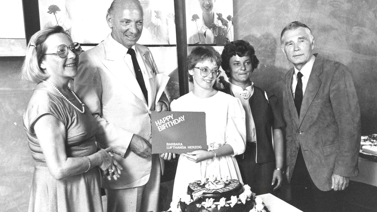 Schwarz-Weiß-Foto einer Gruppe von fünf Personen, die gemeinsam eine Geburtstagskarte über einem Kuchen halten
