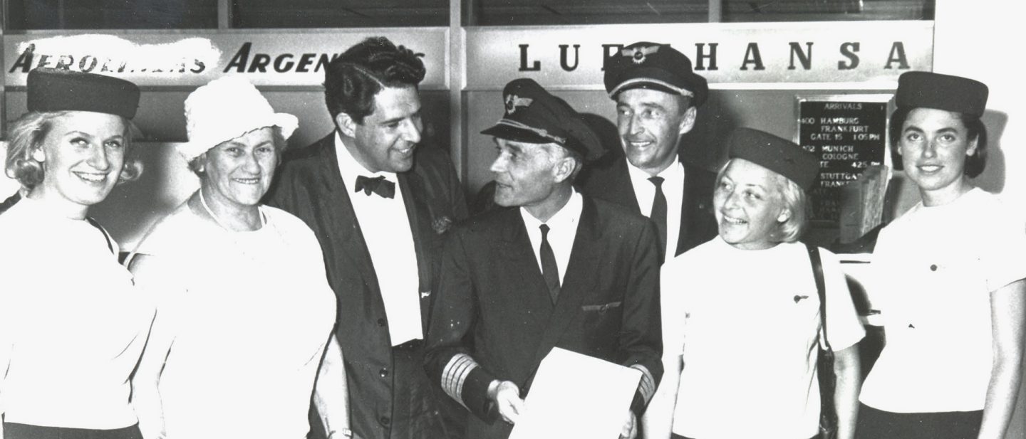 Historisches Schwarz-Weiß-Foto von Lufthansa Piloten und Flugbegleiterinnen, die gemeinsam mit Passagier:innen am Check-in-Schalter stehen