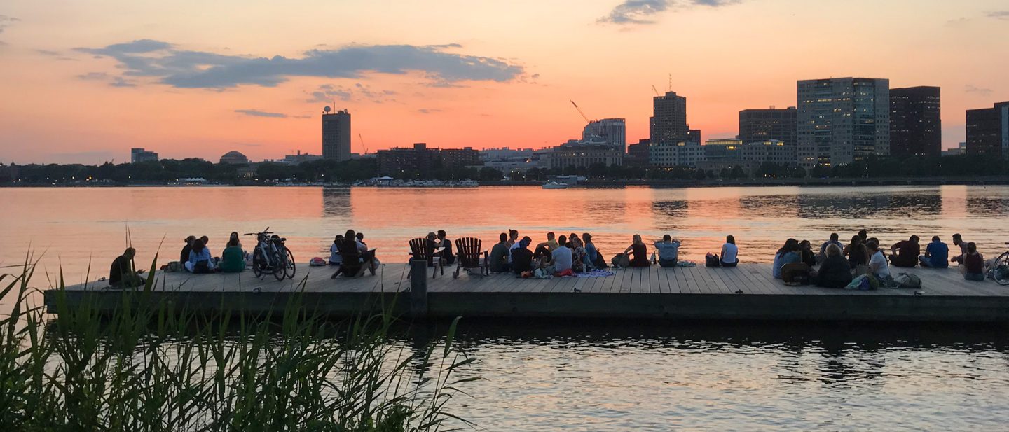 Menschen sitzen bei Sonnenuntergang am Charles River, mit der Bostoner Skyline im Hintergrund