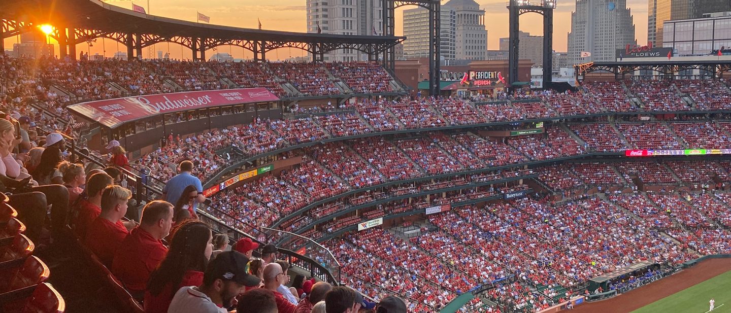 Menge von Fans, die bei Sonnenuntergang ein Baseballspiel im Stadion mit der Skyline der Stadt im Hintergrund beobachtet.