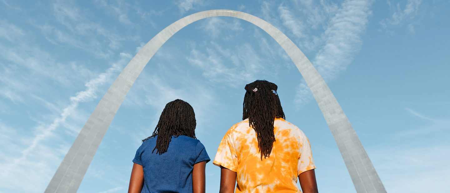 Zwei Personen mit Rastazöpfen stehen vor dem Gateway Arch in St. Louis unter einem blauen Himmel mit leichten Wolken.