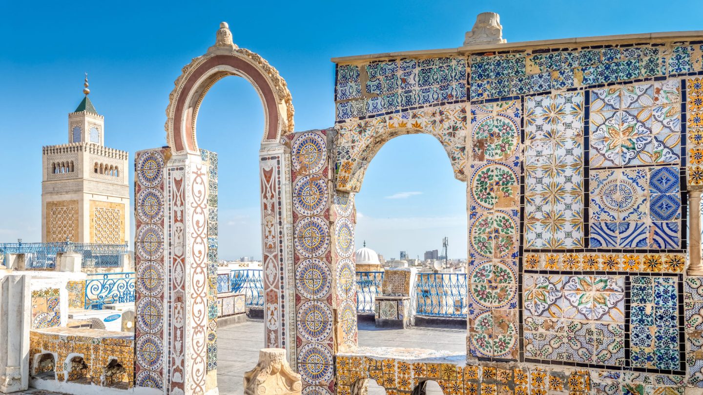 Bunte gekachelte Bögen und ein Balkon mit blauen Geländern vor einem historischen Moscheeturm unter einem klaren Himmel.
