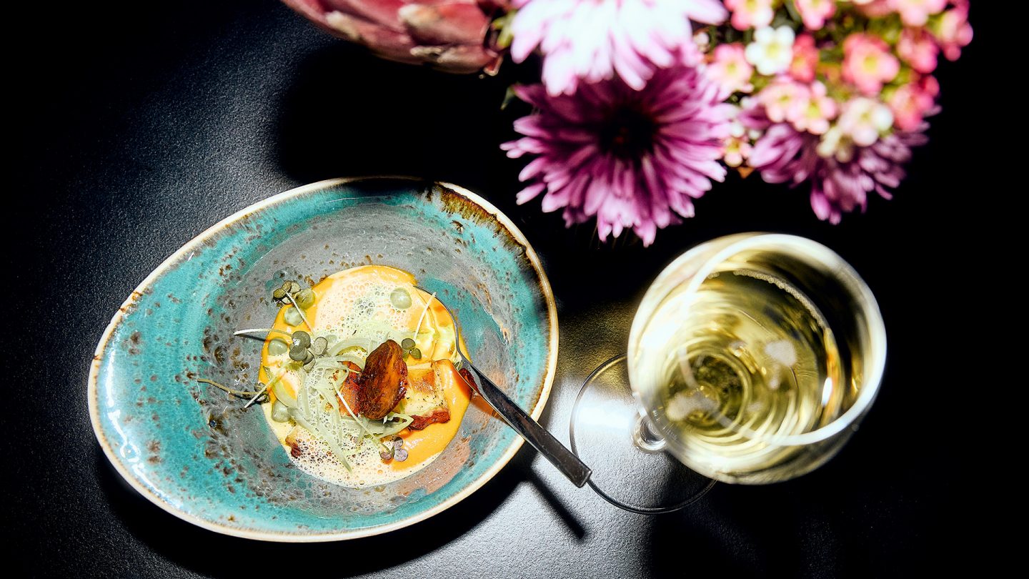 Fein angerichtetes Gourmetgericht in einer Keramikschale neben einem Glas Weißwein und Blumen auf dunkler Tischoberfläche.