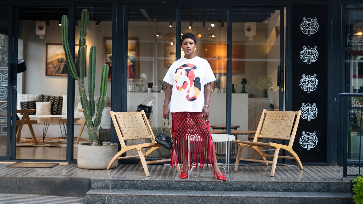 Frau steht vor einem Café mit Glasfront zwischen zwei Stühlen. Sie trägt ein weißes T-Shirt, einen roten Rock und rote Schuhe.