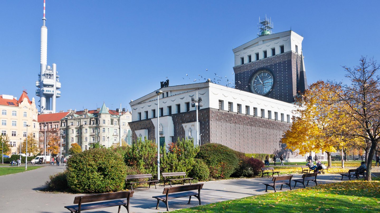 Kirche mit riesiger Uhr im Park, im Hintergrund der Žižkov-Fernsehturm in Vršovice, Prag.