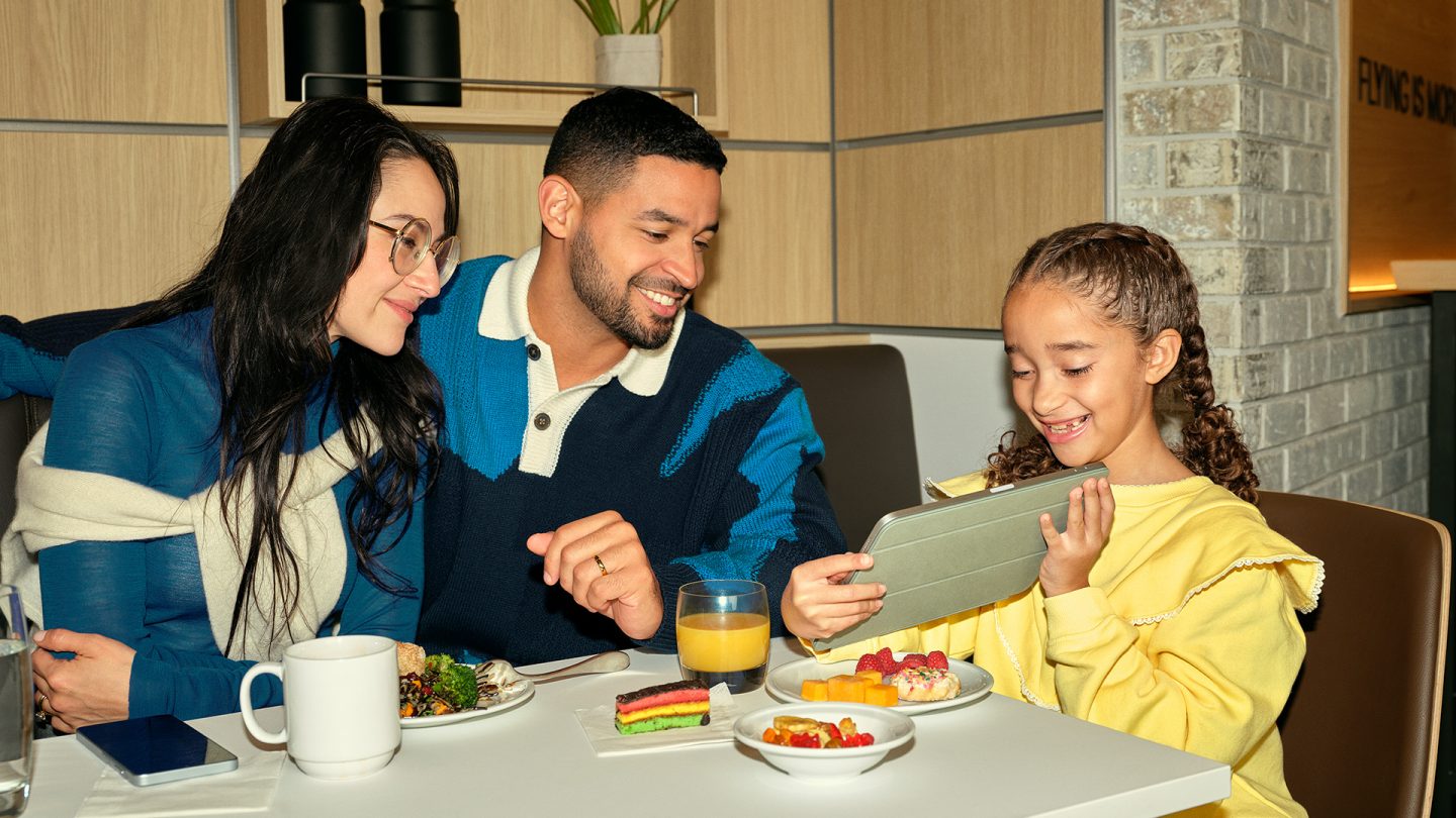 Familie frühstückt in der Lufthansa Lounge, Tochter zeigt etwas am Tablet