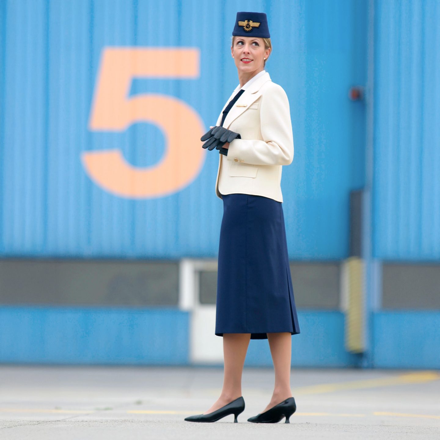 Flugbegleiterin in historischer Lufthansa Uniform mit hellem Blazer, dunklem Rock und Hut mit Abzeichen.