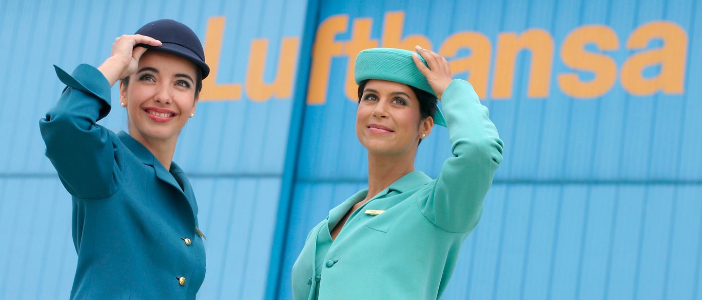 Zwei Lufthansa Flugbegleiterinnen in türkisfarbenen und dunkelblauen Uniformen mit Hüten vor einem Lufthansa Schriftzug.