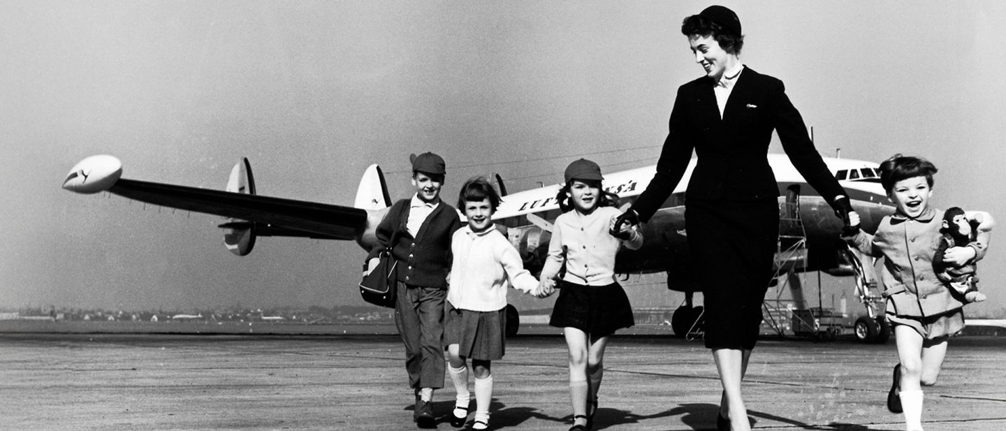 Flugbegleiterin begleitet Kinder über das Rollfeld vor einem Lufthansa Flugzeug.