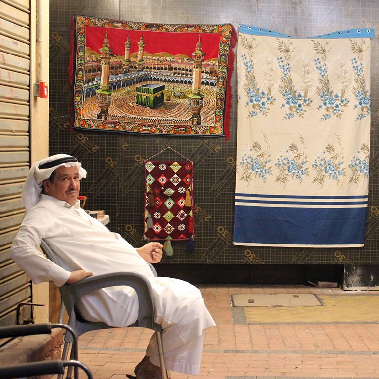 Sitzender Mann in traditioneller Kleidung auf Flohmarkt in Riad vor Wandteppichen mit Kaaba-Motiv und Stoffen