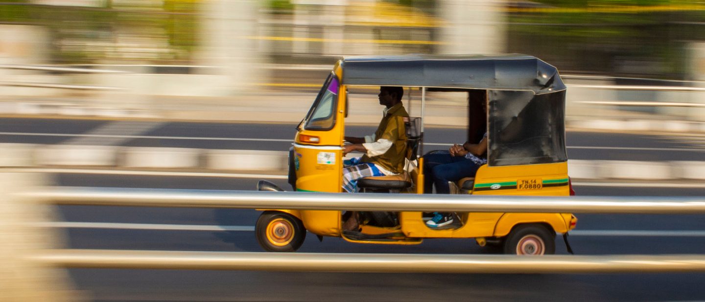 Yellow auto rickshaw with passengers moving fast on a highway with a blurred background.