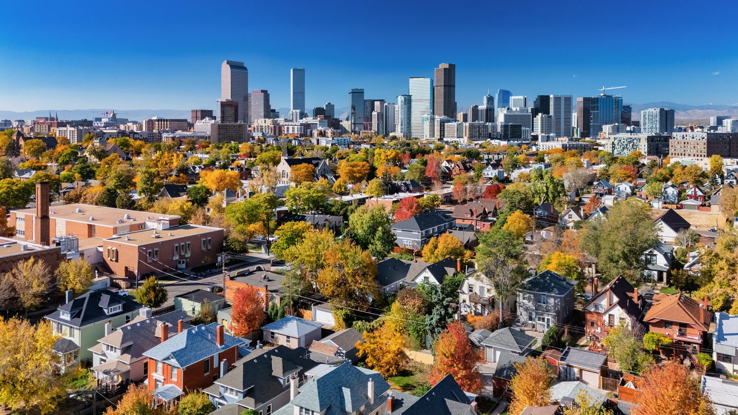 The Denver skyline rises above a neighborhood with colorful fall trees and a clear blue sky