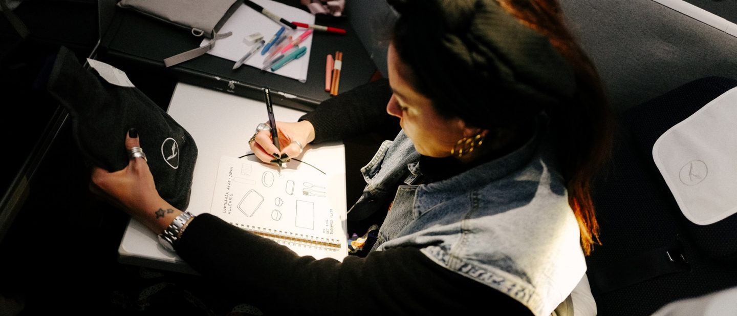 Vue de dessus d'une femme assise à une table dans un avion, dessinant des illustrations dans un carnet de croquis